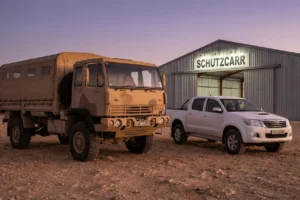 A TCT troop carrier truck parked outside a desert warehouse at dusk, positioned beside a white Toyota Hilux